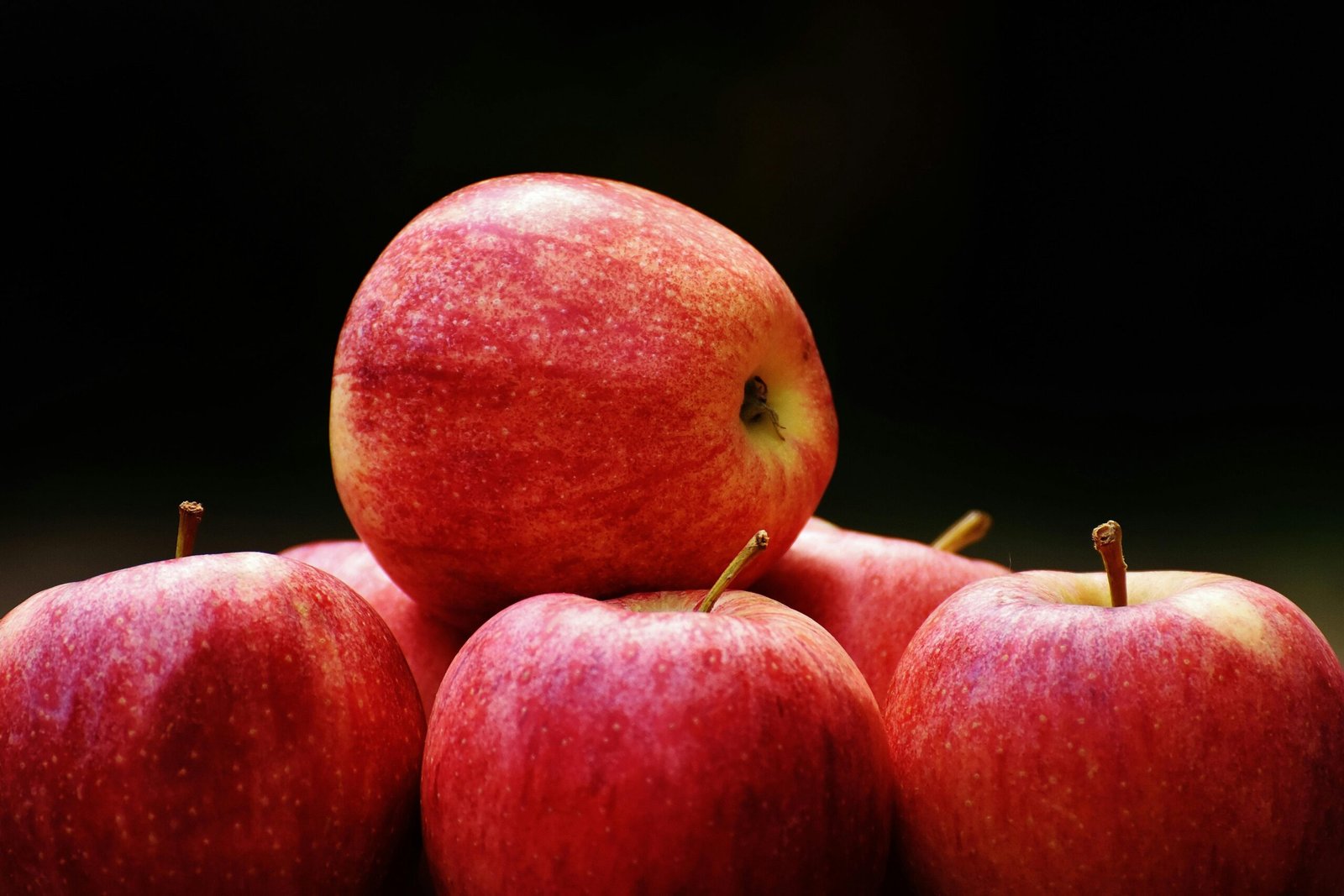 A cluster of ripe, red apples stacked on a dark backdrop highlighting their freshness and vibrancy.