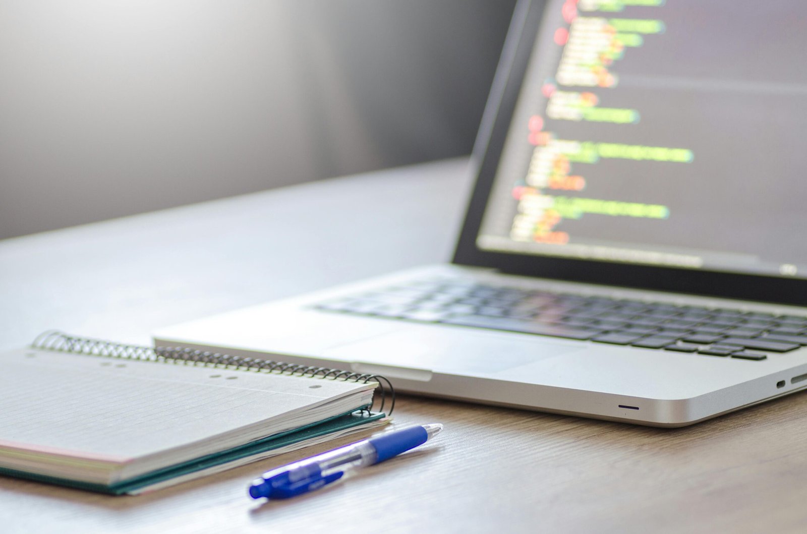 Open laptop with programming code on screen next to a notebook and pen on a desk.